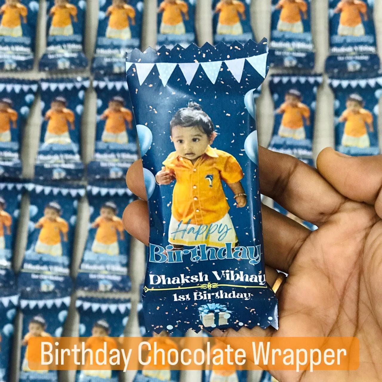 Hand holding a personalized blue chocolate wrapper featuring a baby's photo for a first birthday.