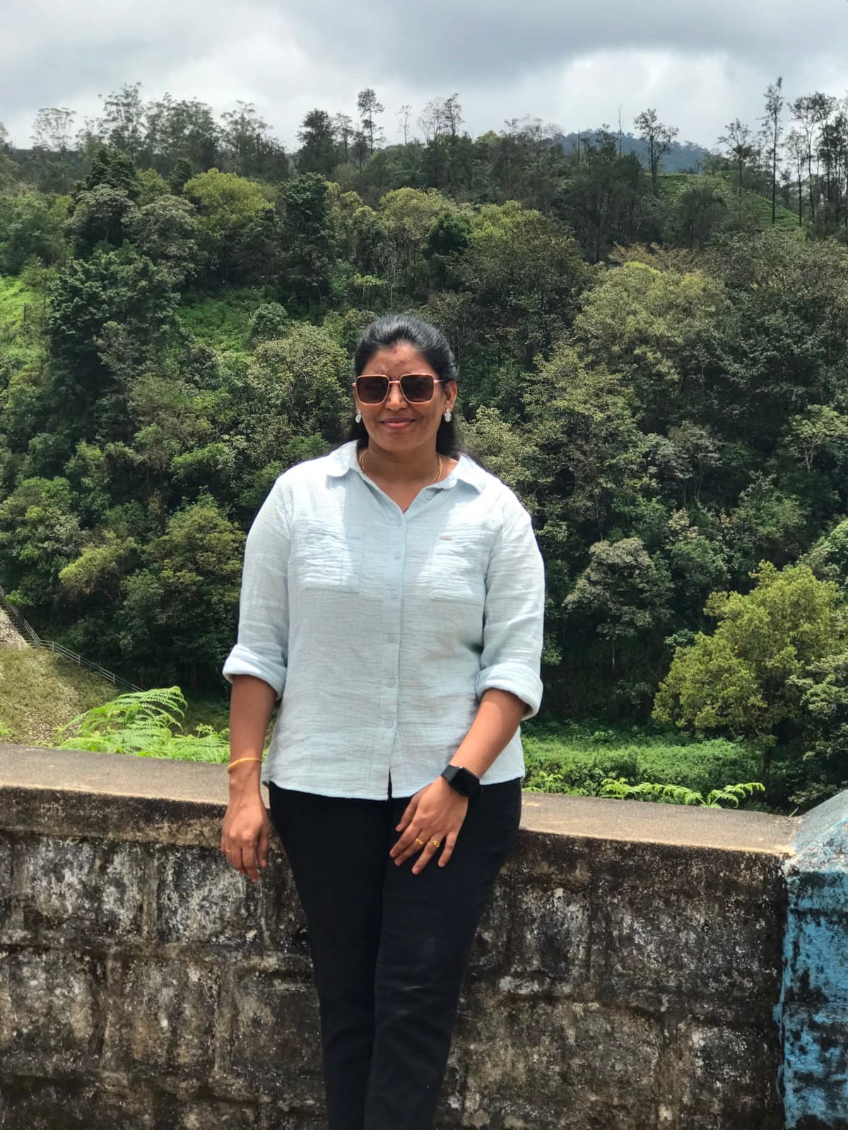 Woman in a light blue shirt and sunglasses stands before a lush, green forest.
