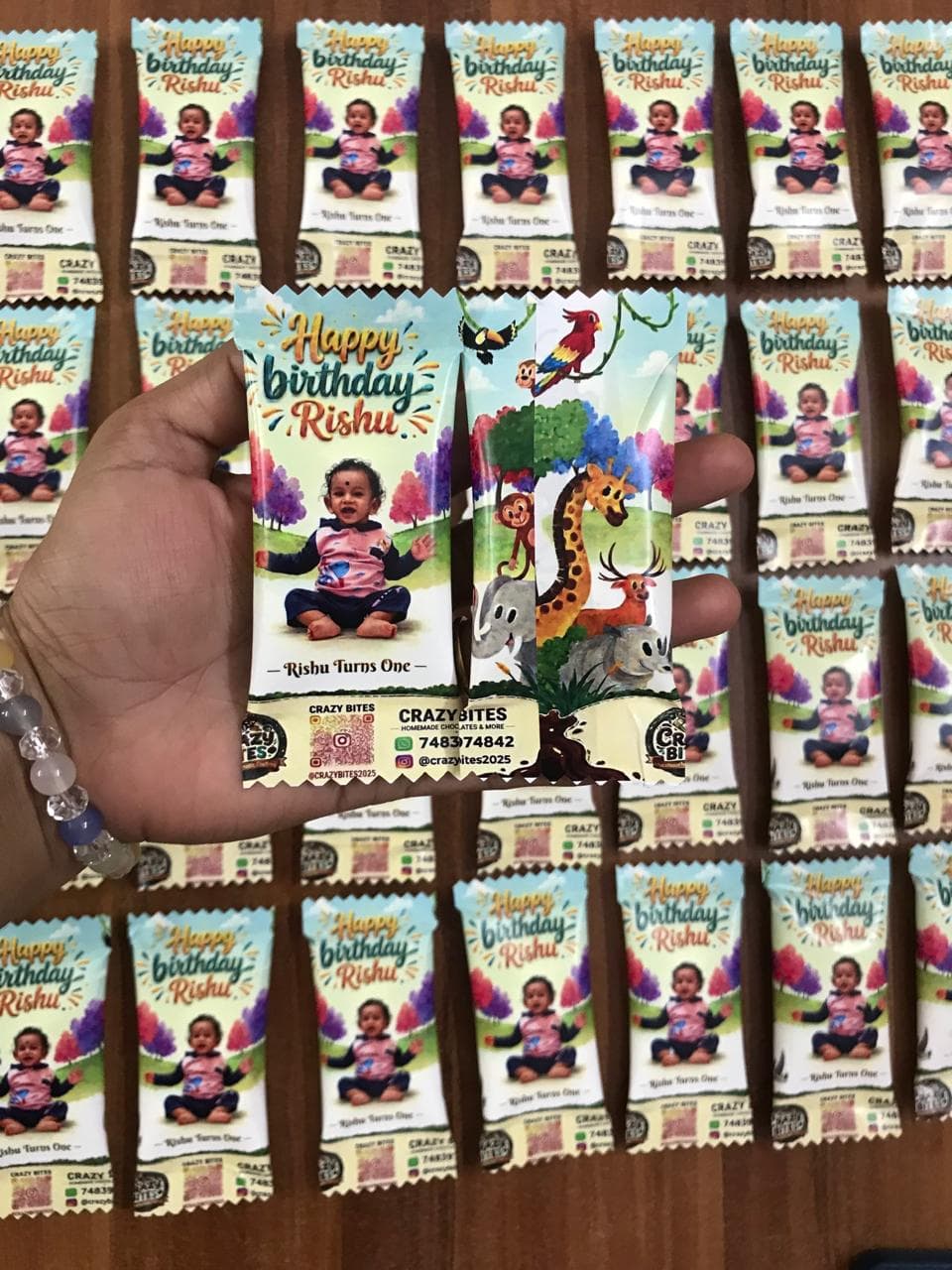 Hand holding personalized first birthday chocolate bars for Rishu featuring a baby and jungle animals.